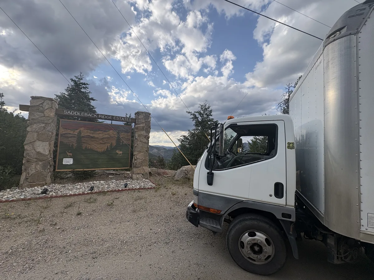 Troops Junk Removal box truck at a stone neighborhood entrance on a gravel mountain road in Evergreen